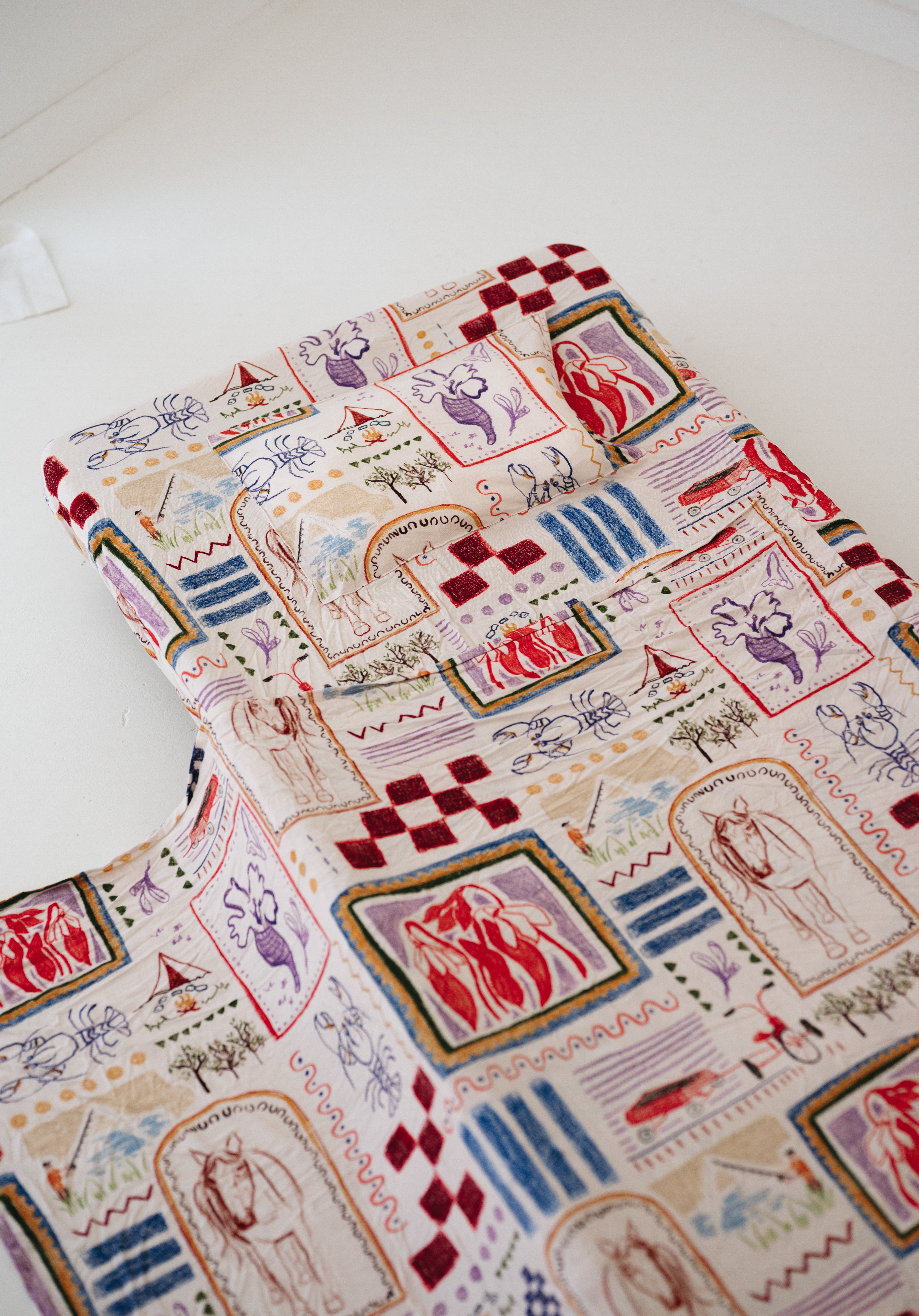 A soft beige and multicolour sheet set laid out onto a daycare stacker bed. The print features hand drawn geometric. The products are photographed with a white background. shapes with nostalgic images including a horse, vintage pedal bike, tent and camp fire, person fishing along with native trees, flowers and animals. Photographed on a white background.