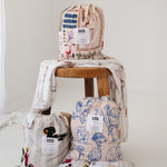 A white and soft grey gingham daycare sheet set packaged in its matching drawstring storage bag drawstring storage bag sitting next to a round wooden stool with alternative daycare sheet set prints available from the same Hello Chooki collection. The print features vintage hand drawn farm animals including chickens, pigs, little girl walking a horse, a boy pushing his dogs in a wheelbarrow, red barn and cows eating hay out of a tractor and trailer. The products are photographed with a white background.
