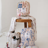 A white and soft grey gingham daycare sheet set packaged in its matching drawstring storage bag drawstring storage bag sitting next to a round wooden stool with alternative daycare sheet set prints available from the same Hello Chooki collection. The print features vintage hand drawn farm animals including chickens, pigs, little girl walking a horse, a boy pushing his dogs in a wheelbarrow, red barn and cows eating hay out of a tractor and trailer. The products are photographed with a white background.