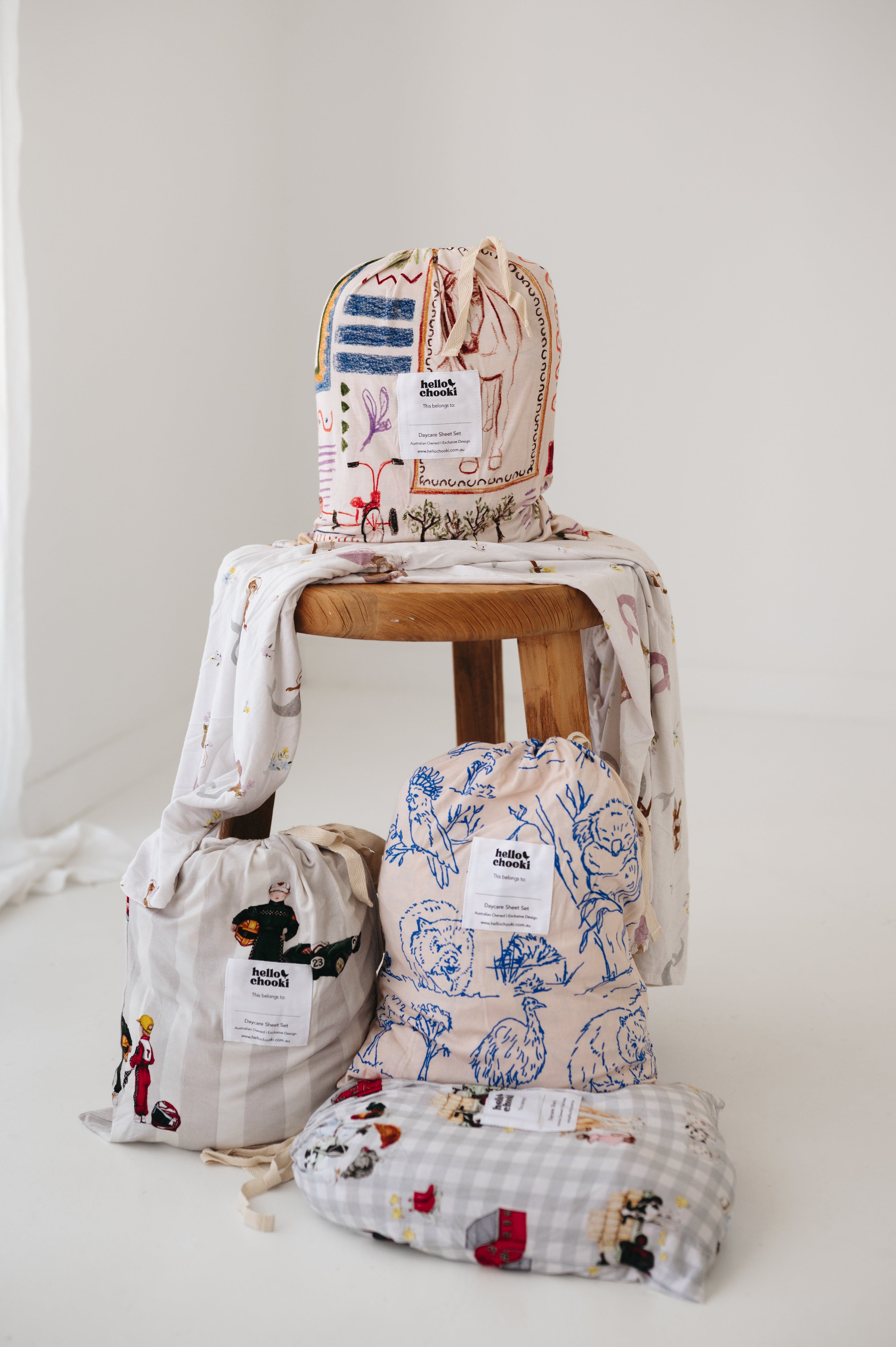 A white and soft grey gingham daycare sheet set packaged in its matching drawstring storage bag drawstring storage bag sitting next to a round wooden stool with alternative daycare sheet set prints available from the same Hello Chooki collection. The print features vintage hand drawn farm animals including chickens, pigs, little girl walking a horse, a boy pushing his dogs in a wheelbarrow, red barn and cows eating hay out of a tractor and trailer. The products are photographed with a white background.