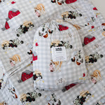 A white and soft grey gingham daycare sheet set packaged in its matching drawstring storage bag drawstring storage bag displayed ontop of the same daycare sheet set laid out onto a daycare stacker bed. The print features vintage hand drawn farm animals including chickens, pigs, little girl walking a horse, a boy pushing his dogs in a wheelbarrow, red barn and cows eating hay out of a tractor and trailer. The products are photographed with a white background.