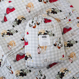 A white and soft grey gingham daycare sheet set packaged in its matching drawstring storage bag drawstring storage bag displayed ontop of the same daycare sheet set laid out onto a daycare stacker bed. The print features vintage hand drawn farm animals including chickens, pigs, little girl walking a horse, a boy pushing his dogs in a wheelbarrow, red barn and cows eating hay out of a tractor and trailer. The products are photographed with a white background.