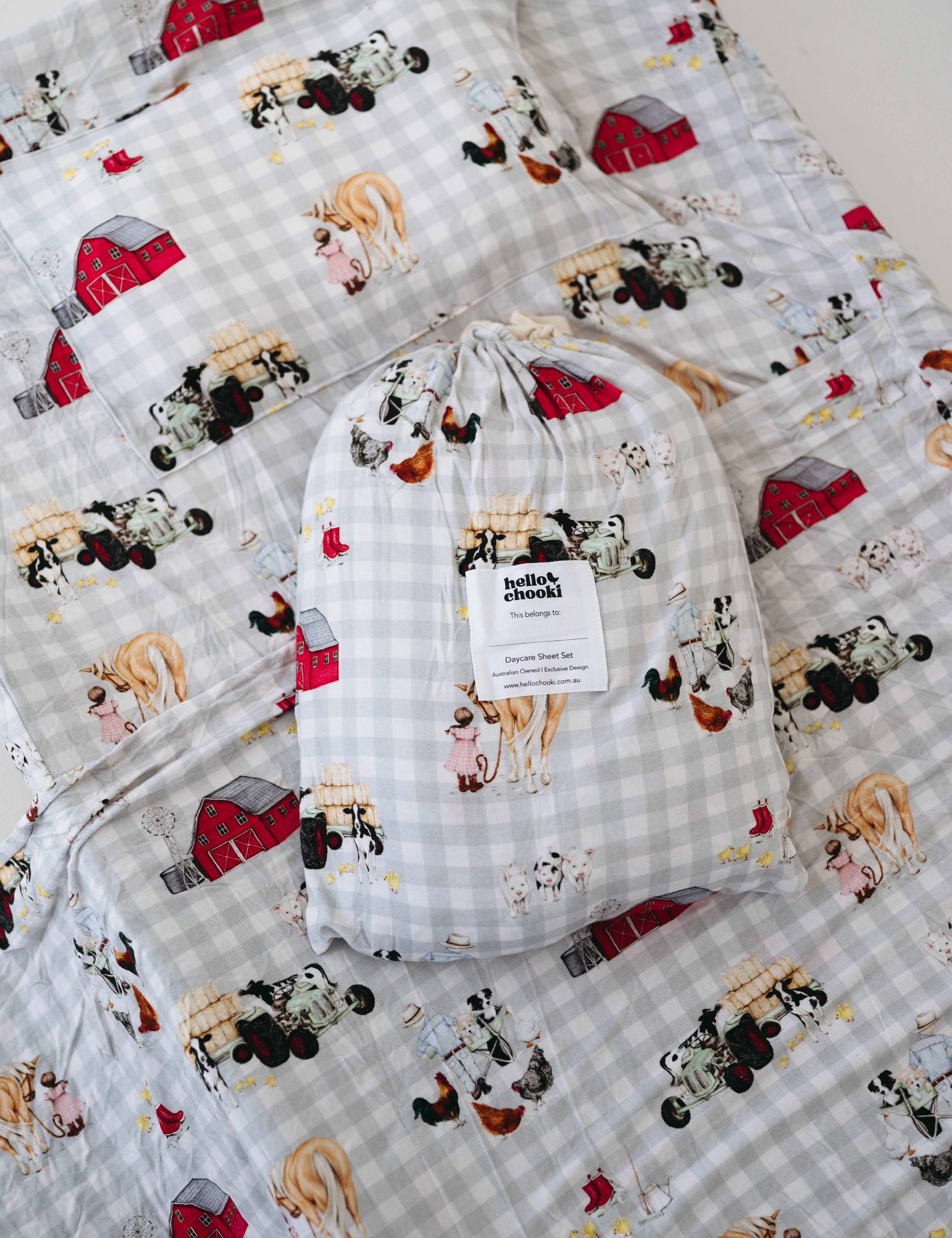 A white and soft grey gingham daycare sheet set packaged in its matching drawstring storage bag drawstring storage bag displayed ontop of the same daycare sheet set laid out onto a daycare stacker bed. The print features vintage hand drawn farm animals including chickens, pigs, little girl walking a horse, a boy pushing his dogs in a wheelbarrow, red barn and cows eating hay out of a tractor and trailer. The products are photographed with a white background.