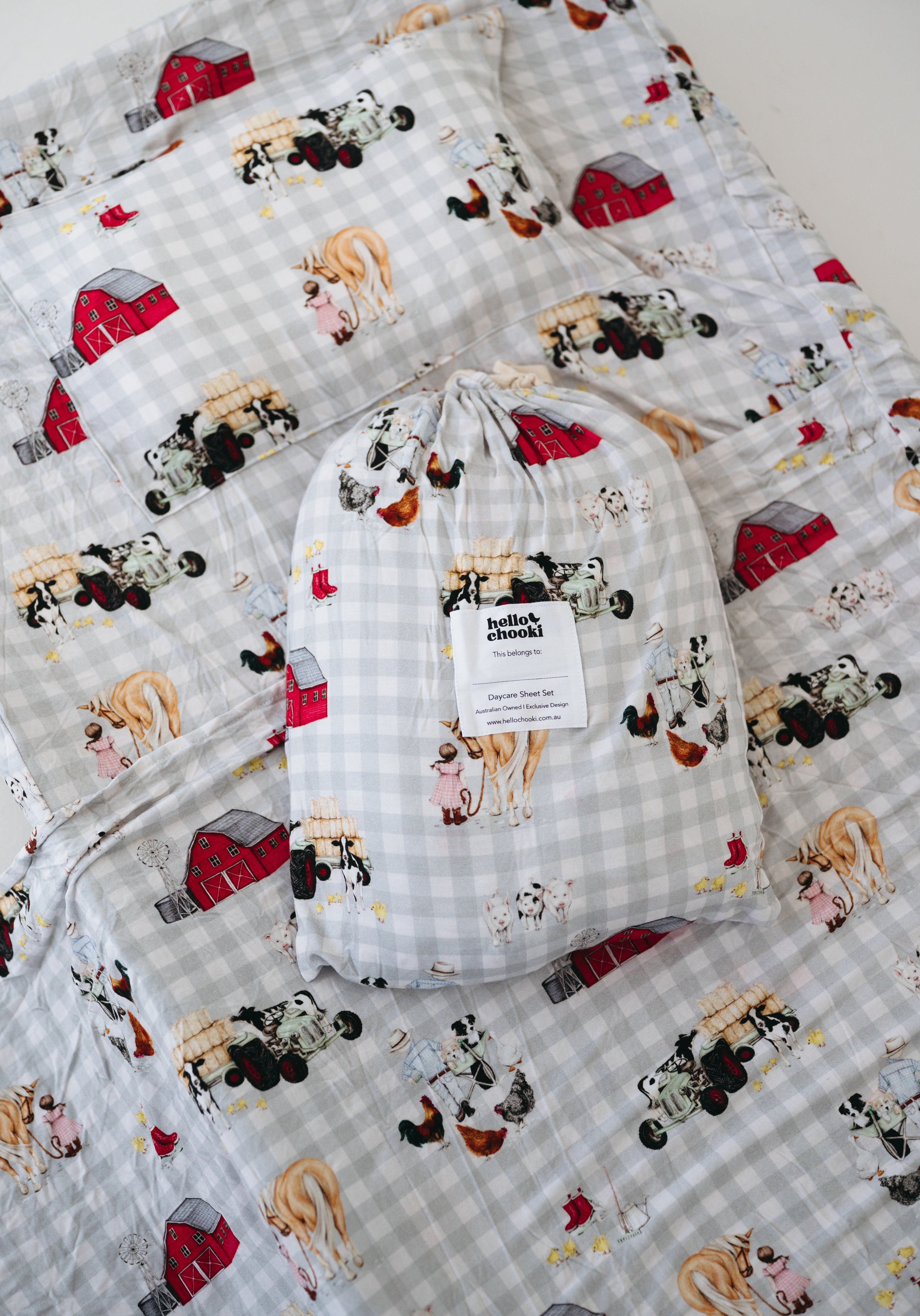 A white and soft grey gingham daycare sheet set packaged in its matching drawstring storage bag drawstring storage bag displayed ontop of the same daycare sheet set laid out onto a daycare stacker bed. The print features vintage hand drawn farm animals including chickens, pigs, little girl walking a horse, a boy pushing his dogs in a wheelbarrow, red barn and cows eating hay out of a tractor and trailer. The products are photographed with a white background.