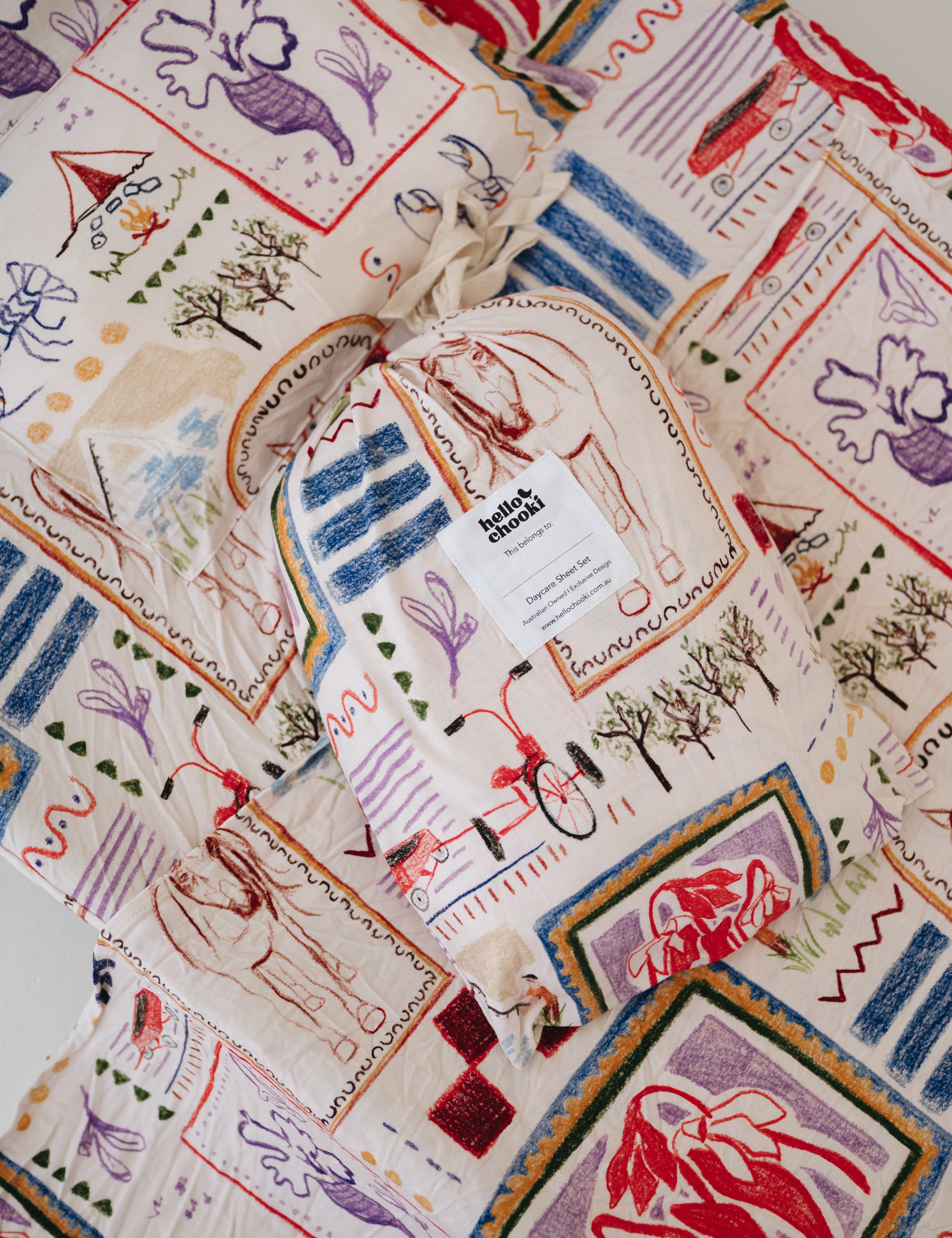 A soft beige and multicolour sheet set packaged in its matching drawstring storage bag displayed ontop of the same daycare sheet set laid out onto a daycare stacker bed. The print features hand drawn geometric. The products are photographed with a white background. shapes with nostalgic images including a horse, vintage pedal bike, tent and camp fire, person fishing along with native trees, flowers and animals. Photographed on a white background.