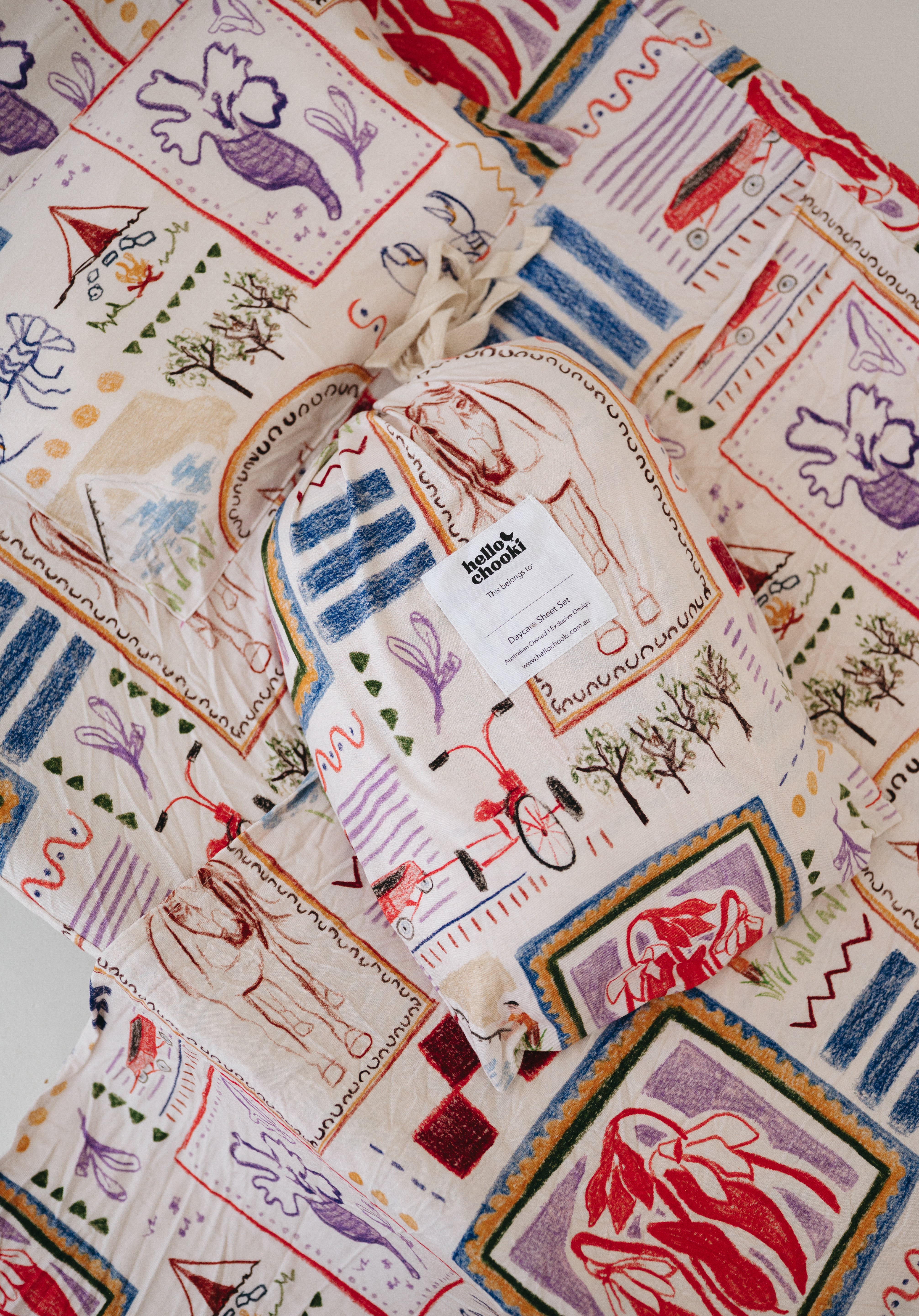A soft beige and multicolour sheet set packaged in its matching drawstring storage bag displayed ontop of the same daycare sheet set laid out onto a daycare stacker bed. The print features hand drawn geometric. The products are photographed with a white background. shapes with nostalgic images including a horse, vintage pedal bike, tent and camp fire, person fishing along with native trees, flowers and animals. Photographed on a white background.
