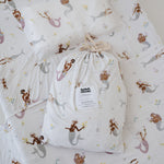 A white and multicolour bohemian style mermaid sheet set packaged in its matching drawstring storage bag displayed on the same daycare sheet set laid out onto a daycare stacker bed. The print features hand painted mermaids of different tones, with various colour tails and hair colours including soft blue, soft purple and soft pink. The mermaids have flowers in their hair and hands with same flowers scattered throughout. The products are photographed with a white background.