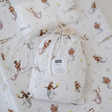 A white and multicolour bohemian style mermaid sheet set packaged in its matching drawstring storage bag displayed on the same daycare sheet set laid out onto a daycare stacker bed. The print features hand painted mermaids of different tones, with various colour tails and hair colours including soft blue, soft purple and soft pink. The mermaids have flowers in their hair and hands with same flowers scattered throughout. The products are photographed with a white background.