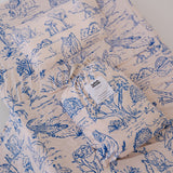 A soft beige and blue daycare sheet set packaged in its matching drawstring storage bag displayed ontop of the same daycare sheet set laid out onto a daycare stacker bed. The print features hand drawn outback Australian animals and natives including emu's, crocodiles, kangaroos, koala's, wombats, echidna's with native gum trees and plants. The products are photographed with a white background.