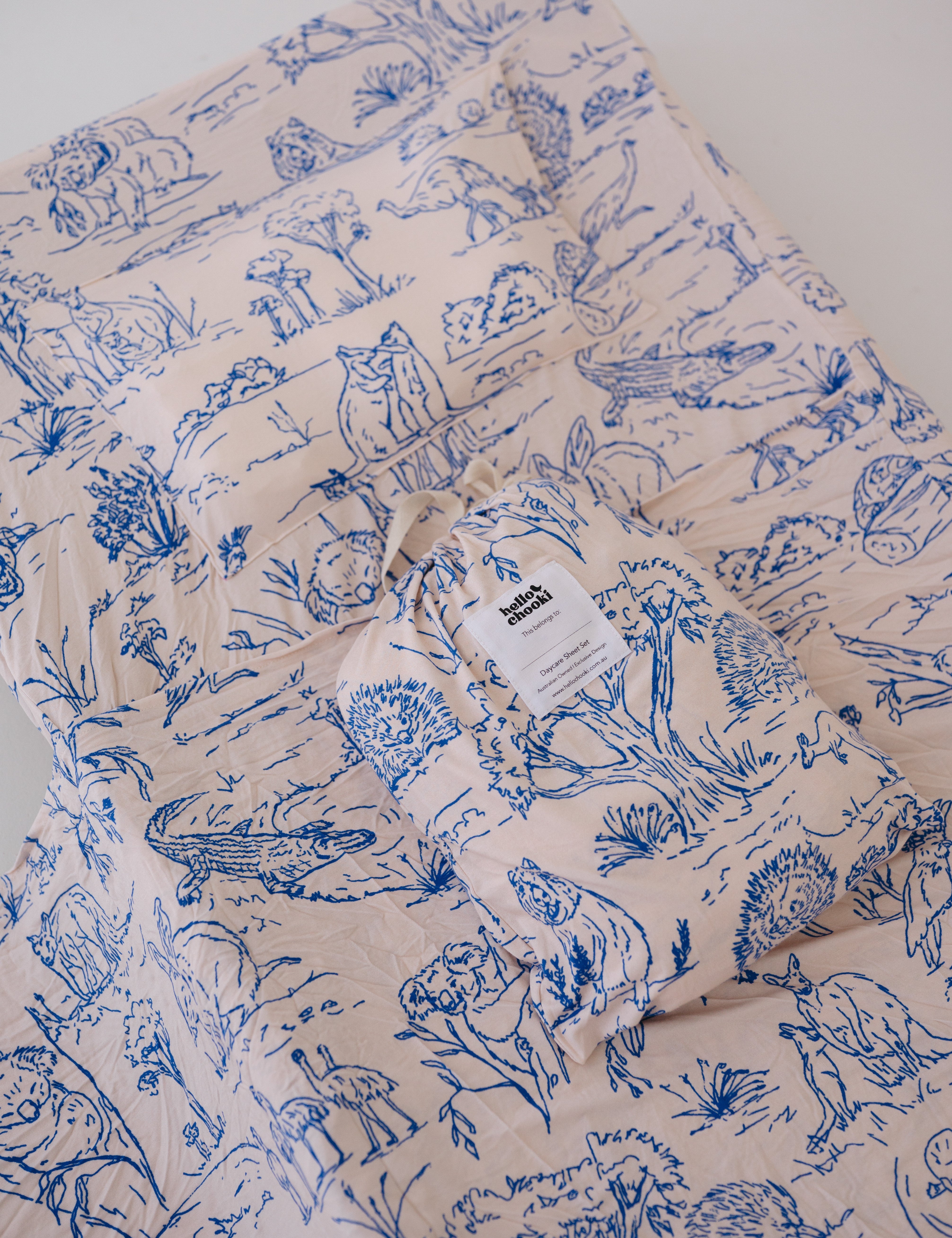 A soft beige and blue daycare sheet set packaged in its matching drawstring storage bag displayed ontop of the same daycare sheet set laid out onto a daycare stacker bed. The print features hand drawn outback Australian animals and natives including emu's, crocodiles, kangaroos, koala's, wombats, echidna's with native gum trees and plants. The products are photographed with a white background.