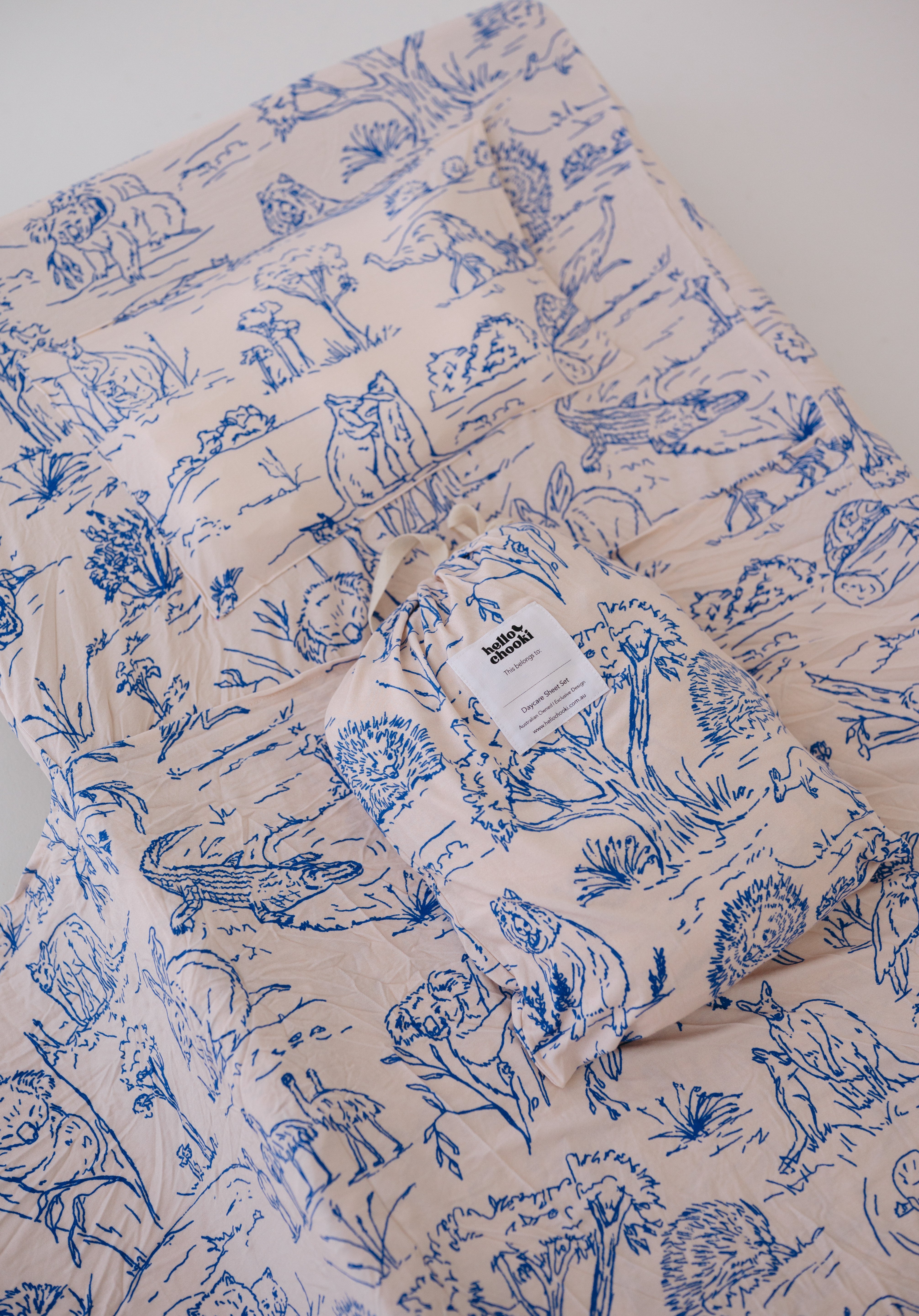 A soft beige and blue daycare sheet set packaged in its matching drawstring storage bag displayed ontop of the same daycare sheet set laid out onto a daycare stacker bed. The print features hand drawn outback Australian animals and natives including emu's, crocodiles, kangaroos, koala's, wombats, echidna's with native gum trees and plants. The products are photographed with a white background.