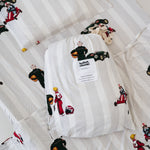 A white and soft grey striped daycare sheet set packaged in its matching drawstring storage bag drawstring storage bag placed onto of the same daycare sheet set laid out onto a daycare stacker bed. The print features vintage hand drawn boys dressed in vintage race overalls and helmet next to a vintage race car and driving a vintage go kart. The products are photographed with a white background.