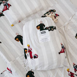 A white and soft grey striped daycare sheet set packaged in its matching drawstring storage bag drawstring storage bag placed onto of the same daycare sheet set laid out onto a daycare stacker bed. The print features vintage hand drawn boys dressed in vintage race overalls and helmet next to a vintage race car and driving a vintage go kart. The products are photographed with a white background.