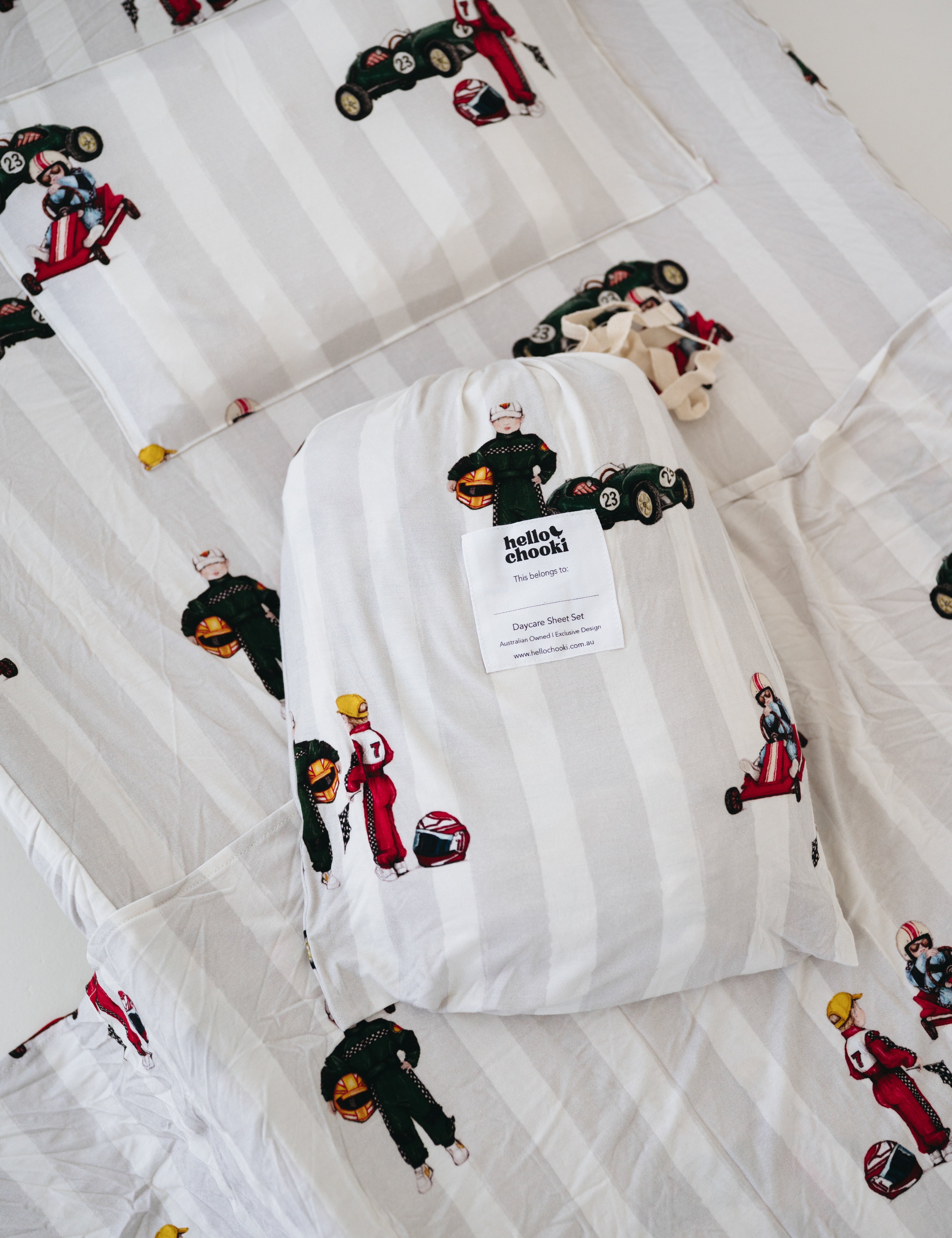 A white and soft grey striped daycare sheet set packaged in its matching drawstring storage bag drawstring storage bag placed onto of the same daycare sheet set laid out onto a daycare stacker bed. The print features vintage hand drawn boys dressed in vintage race overalls and helmet next to a vintage race car and driving a vintage go kart. The products are photographed with a white background.