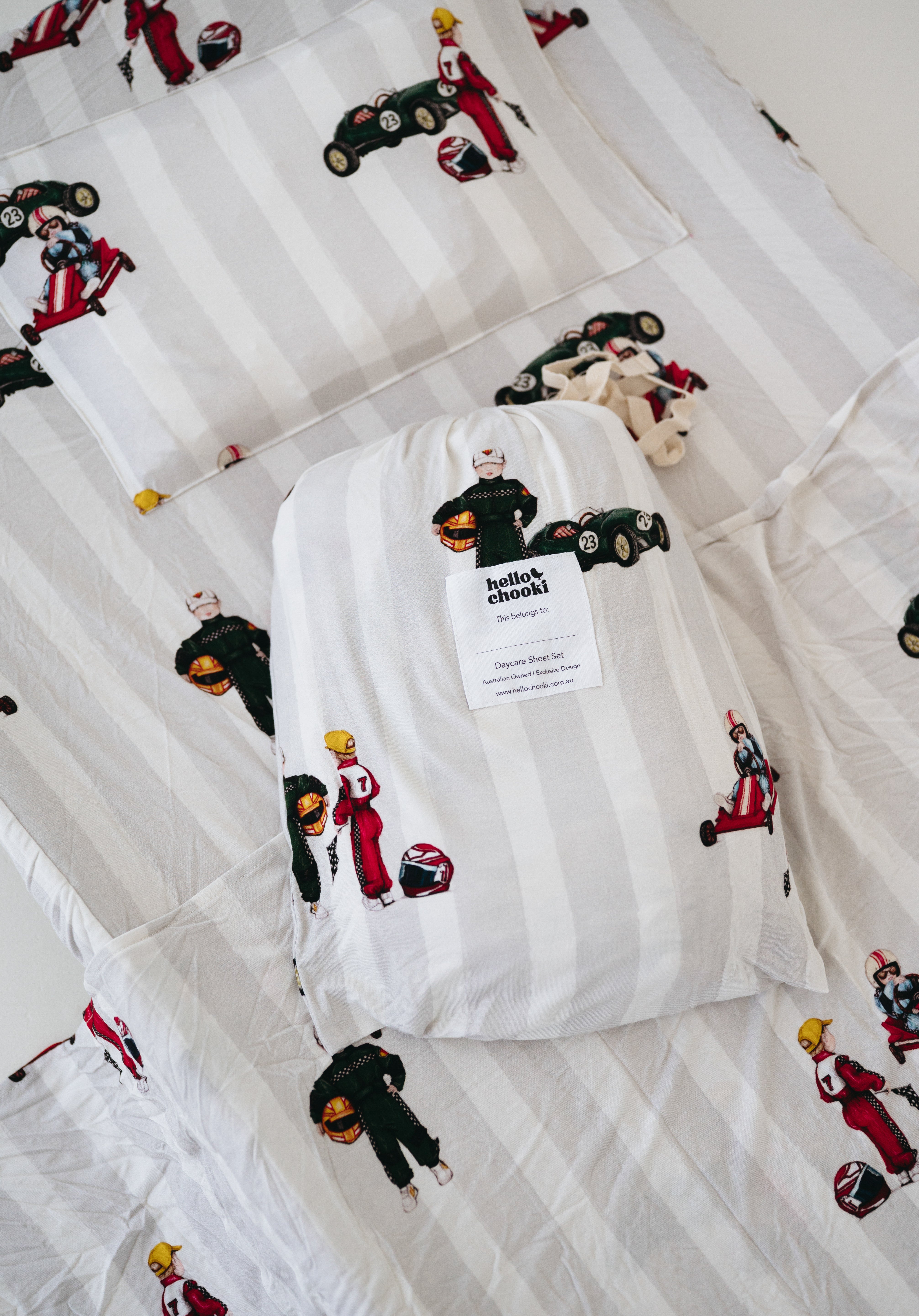 A white and soft grey striped daycare sheet set packaged in its matching drawstring storage bag drawstring storage bag placed onto of the same daycare sheet set laid out onto a daycare stacker bed. The print features vintage hand drawn boys dressed in vintage race overalls and helmet next to a vintage race car and driving a vintage go kart. The products are photographed with a white background.