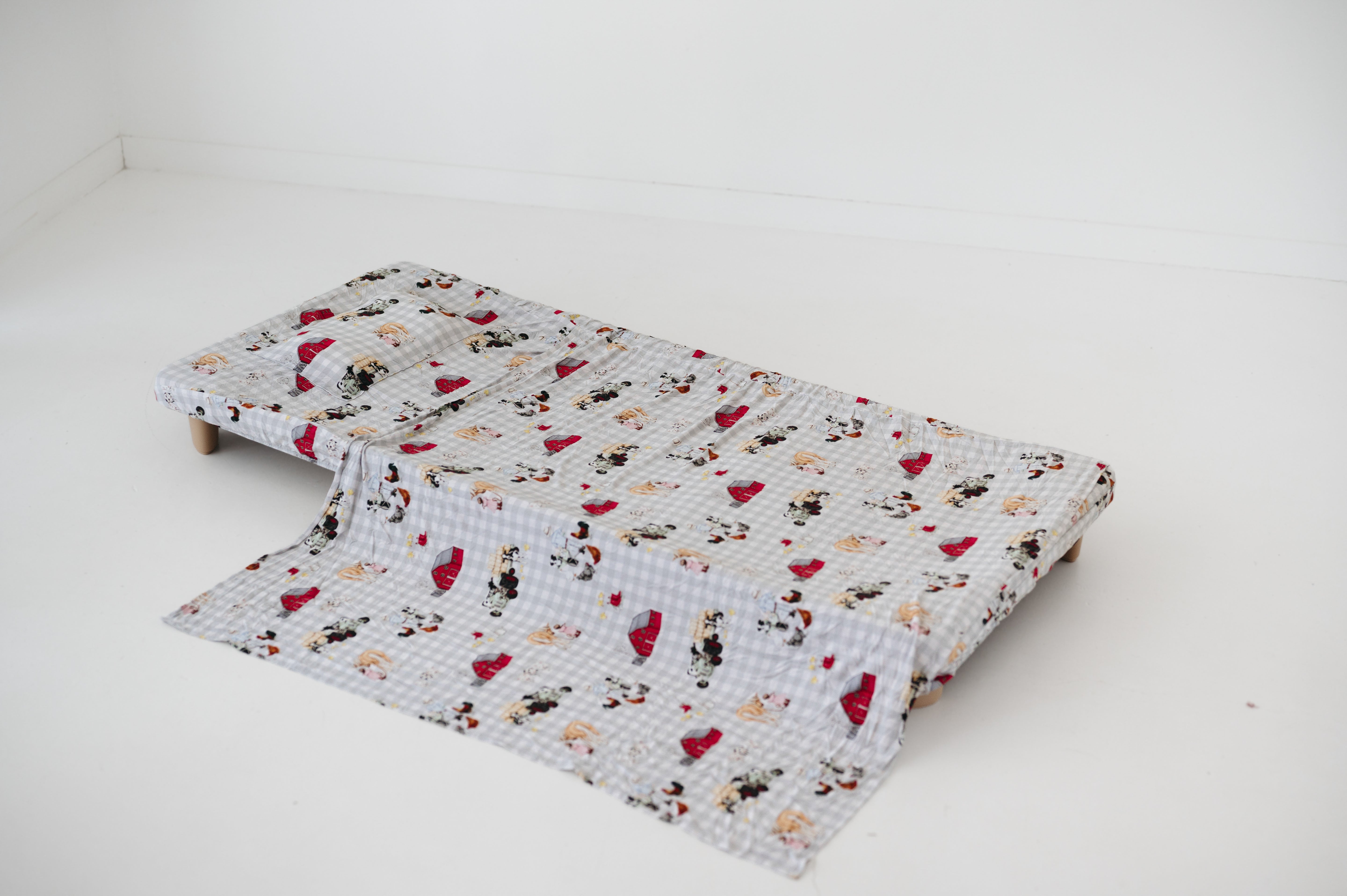 A white and soft grey gingham daycare sheet set laid out onto a daycare stacker bed. The print features vintage hand drawn farm animals including chickens, pigs, little girl walking a horse, a boy pushing his dogs in a wheelbarrow, red barn and cows eating hay out of a tractor and trailer. The products are photographed with a white background.