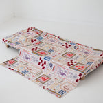 A soft beige and multicolour sheet set laid out onto a daycare stacker bed. The print features hand drawn geometric. The products are photographed with a white background. shapes with nostalgic images including a horse, vintage pedal bike, tent and camp fire, person fishing along with native trees, flowers and animals. Photographed on a white background.