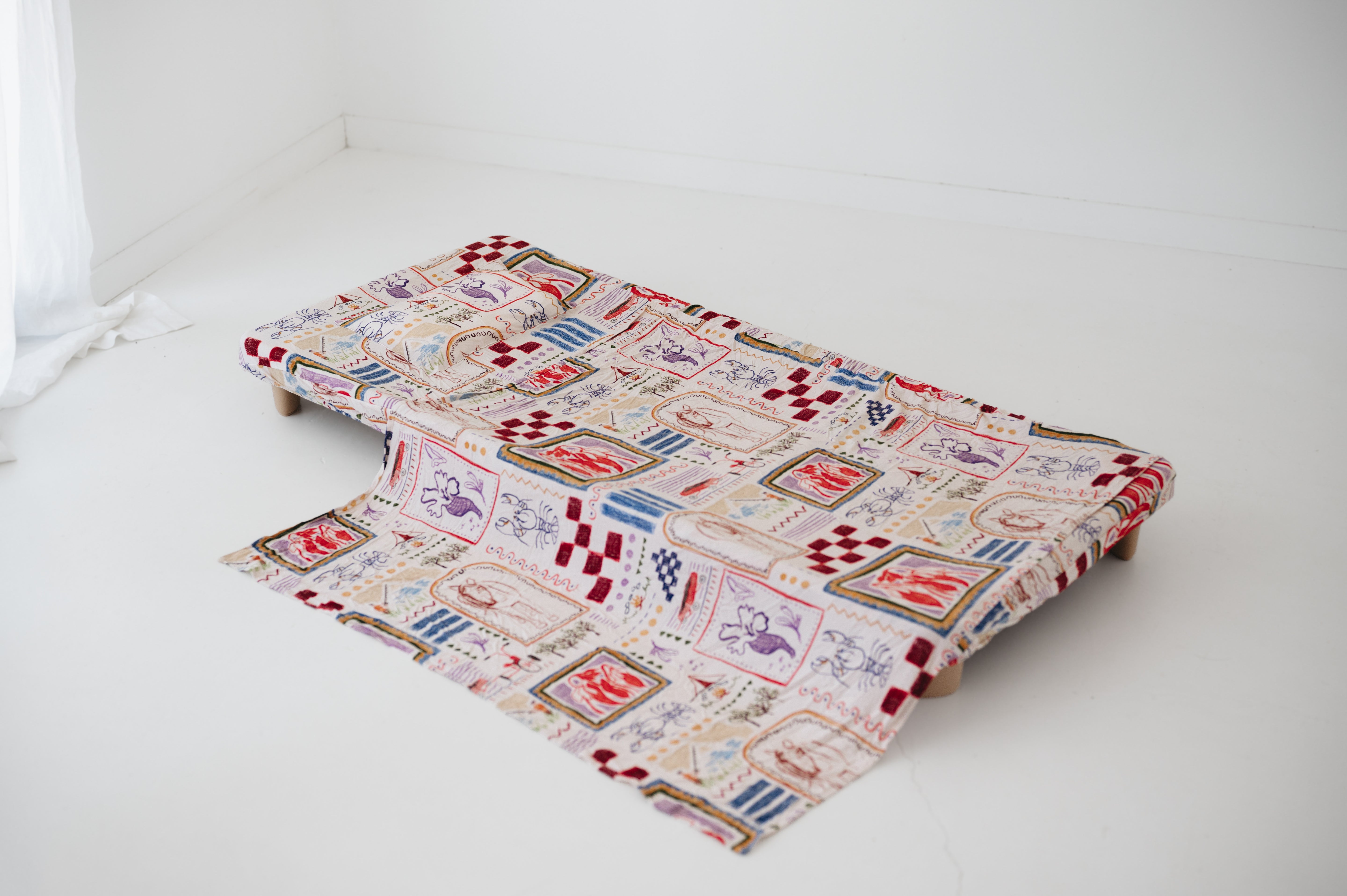 A soft beige and multicolour sheet set laid out onto a daycare stacker bed. The print features hand drawn geometric. The products are photographed with a white background. shapes with nostalgic images including a horse, vintage pedal bike, tent and camp fire, person fishing along with native trees, flowers and animals. Photographed on a white background.