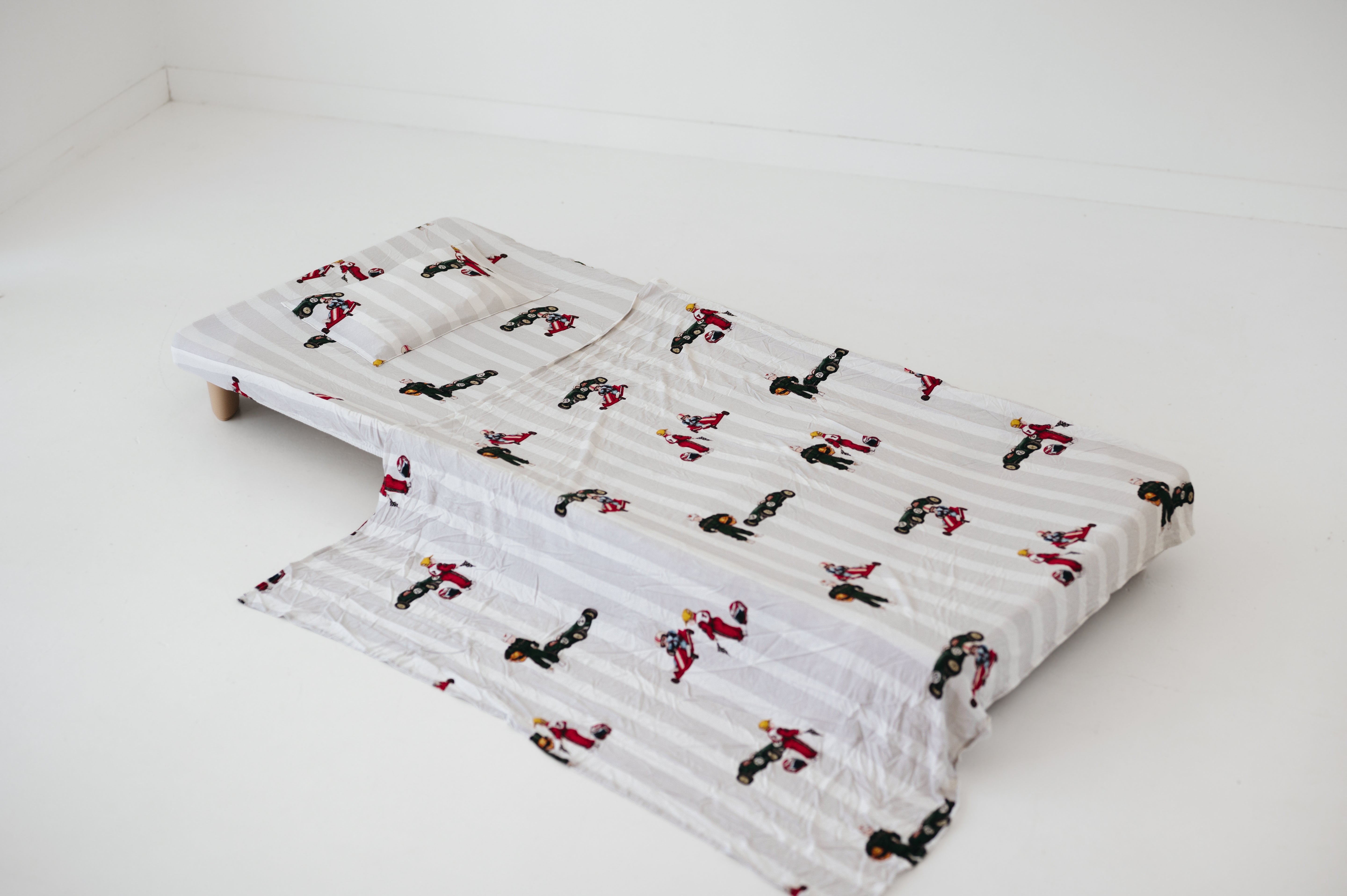A white and soft grey striped daycare sheet set laid out onto a daycare stacker bed. The print features vintage hand drawn boys dressed in vintage race overalls and helmet next to a vintage race car and driving a vintage go kart. The products are photographed with a white background.