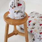 A white and soft grey gingham daycare sheet set packaged in its matching drawstring storage bag drawstring storage bag sitting on a round wooden stool next to the same daycare sheet set laid out onto a daycare stacker bed. The print features vintage hand drawn farm animals including chickens, pigs, little girl walking a horse, a boy pushing his dogs in a wheelbarrow, red barn and cows eating hay out of a tractor and trailer. The products are photographed with a white background.