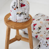 A white and soft grey gingham daycare sheet set packaged in its matching drawstring storage bag drawstring storage bag sitting on a round wooden stool next to the same daycare sheet set laid out onto a daycare stacker bed. The print features vintage hand drawn farm animals including chickens, pigs, little girl walking a horse, a boy pushing his dogs in a wheelbarrow, red barn and cows eating hay out of a tractor and trailer. The products are photographed with a white background.