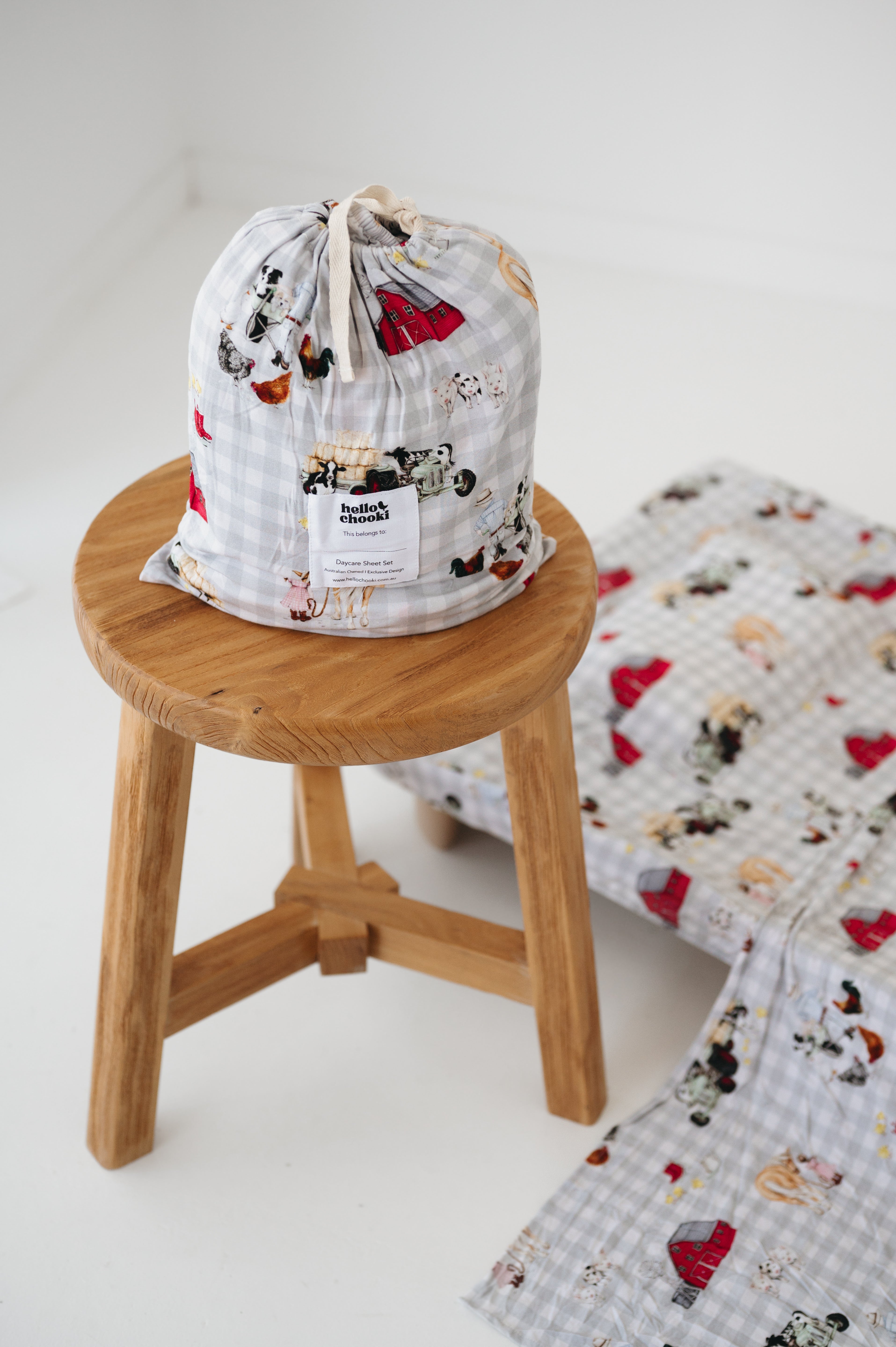 A white and soft grey gingham daycare sheet set packaged in its matching drawstring storage bag drawstring storage bag sitting on a round wooden stool next to the same daycare sheet set laid out onto a daycare stacker bed. The print features vintage hand drawn farm animals including chickens, pigs, little girl walking a horse, a boy pushing his dogs in a wheelbarrow, red barn and cows eating hay out of a tractor and trailer. The products are photographed with a white background.