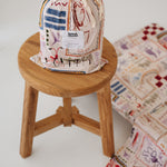 A soft beige and multicolour sheet set packaged in its matching drawstring storage bag sitting on a round wooden stool next to the same daycare sheet set laid out onto a daycare stacker bed. The print features hand drawn geometric. The products are photographed with a white background. shapes with nostalgic images including a horse, vintage pedal bike, tent and camp fire, person fishing along with native trees, flowers and animals. Photographed on a white background.