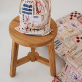 A soft beige and multicolour sheet set packaged in its matching drawstring storage bag sitting on a round wooden stool next to the same daycare sheet set laid out onto a daycare stacker bed. The print features hand drawn geometric. The products are photographed with a white background. shapes with nostalgic images including a horse, vintage pedal bike, tent and camp fire, person fishing along with native trees, flowers and animals. Photographed on a white background.
