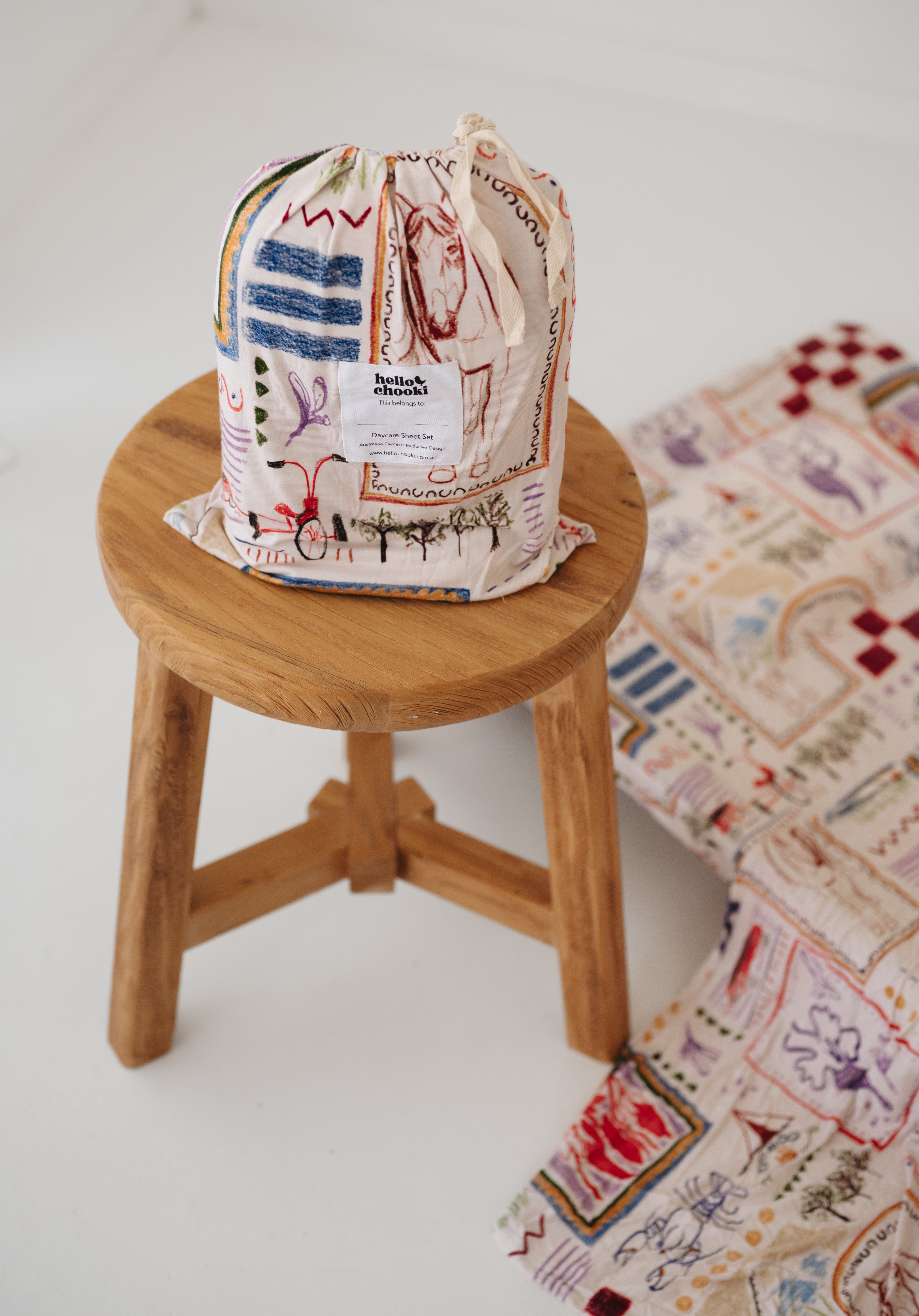 A soft beige and multicolour sheet set packaged in its matching drawstring storage bag sitting on a round wooden stool next to the same daycare sheet set laid out onto a daycare stacker bed. The print features hand drawn geometric. The products are photographed with a white background. shapes with nostalgic images including a horse, vintage pedal bike, tent and camp fire, person fishing along with native trees, flowers and animals. Photographed on a white background.