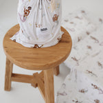 A white and multicolour bohemian style mermaid sheet set packaged in its matching drawstring storage bag sitting on a round wooden stool next to the same daycare sheet set laid out onto a daycare stacker bed. The print features hand painted mermaids of different tones, with various colour tails and hair colours including soft blue, soft purple and soft pink. The mermaids have flowers in their hair and hands with same flowers scattered throughout. The products are photographed with a white background.