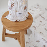 A white and multicolour bohemian style mermaid sheet set packaged in its matching drawstring storage bag sitting on a round wooden stool next to the same daycare sheet set laid out onto a daycare stacker bed. The print features hand painted mermaids of different tones, with various colour tails and hair colours including soft blue, soft purple and soft pink. The mermaids have flowers in their hair and hands with same flowers scattered throughout. The products are photographed with a white background.