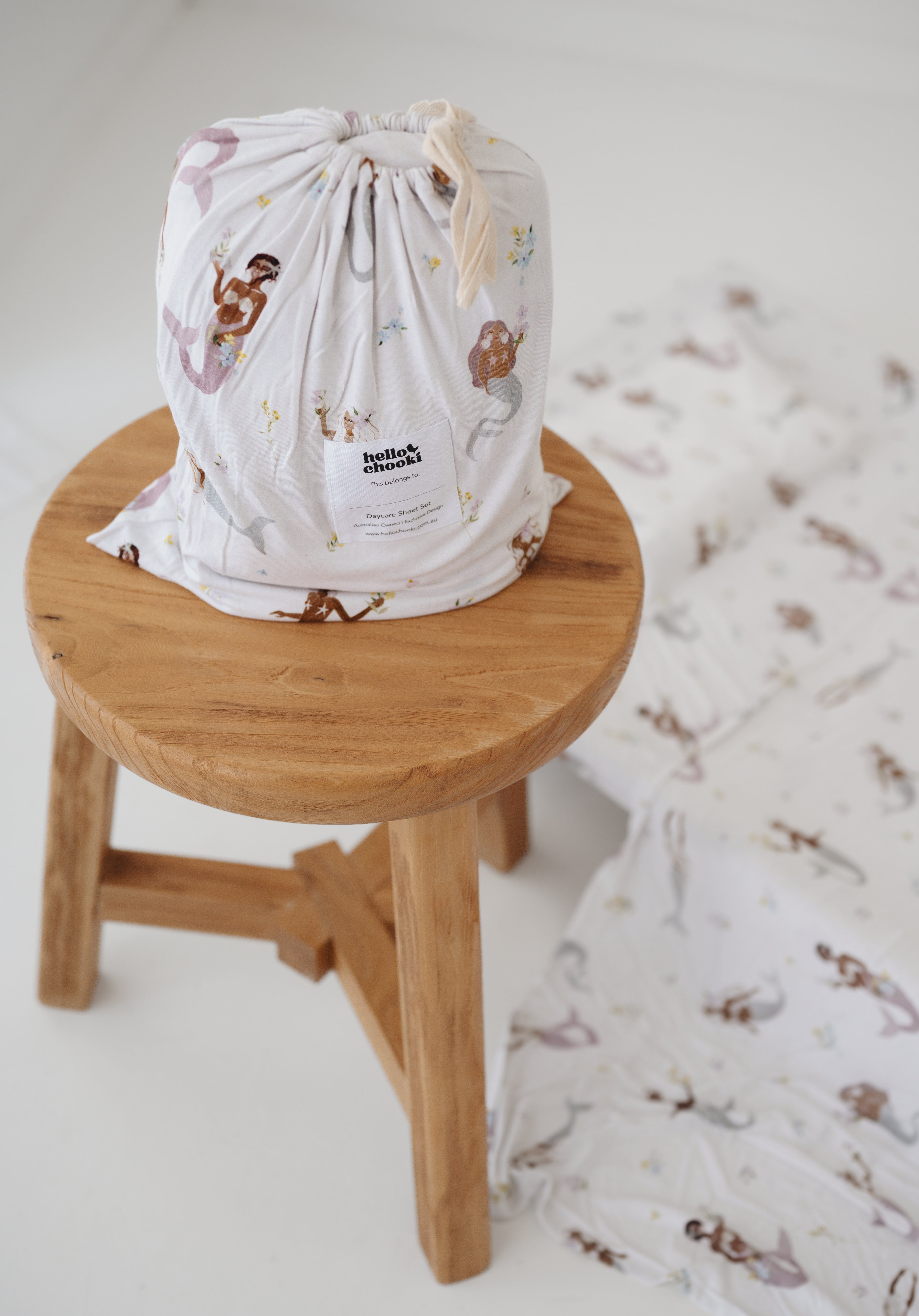 A white and multicolour bohemian style mermaid sheet set packaged in its matching drawstring storage bag sitting on a round wooden stool next to the same daycare sheet set laid out onto a daycare stacker bed. The print features hand painted mermaids of different tones, with various colour tails and hair colours including soft blue, soft purple and soft pink. The mermaids have flowers in their hair and hands with same flowers scattered throughout. The products are photographed with a white background.