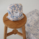 A soft beige and blue daycare sheet set packaged in its matching drawstring storage bag sitting on a round wooden stool next to the same daycare sheet set laid out onto a daycare stacker bed. The print features hand drawn outback Australian animals and natives including emu's, crocodiles, kangaroos, koala's, wombats, echidna's with native gum trees and plants. The products are photographed with a white background.