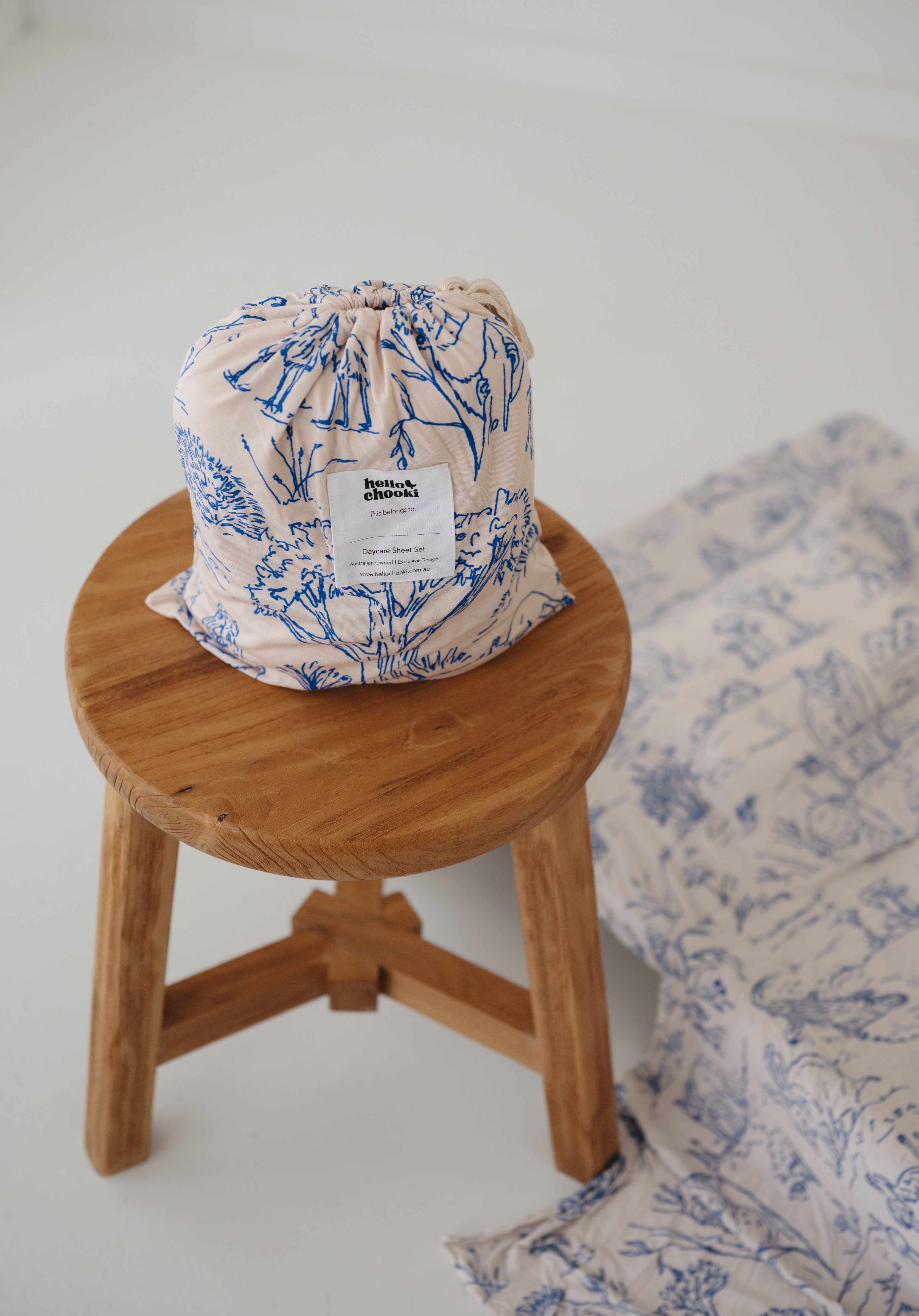 A soft beige and blue daycare sheet set packaged in its matching drawstring storage bag sitting on a round wooden stool next to the same daycare sheet set laid out onto a daycare stacker bed. The print features hand drawn outback Australian animals and natives including emu's, crocodiles, kangaroos, koala's, wombats, echidna's with native gum trees and plants. The products are photographed with a white background.