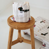 A white and soft grey striped daycare sheet set packaged in its matching drawstring storage bag drawstring storage bag sitting on a round wooden stool next to the same daycare sheet set laid out onto a daycare stacker bed. The print features vintage hand drawn boys dressed in vintage race overalls and helmet next to a vintage race car and driving a vintage go kart. The products are photographed with a white background.