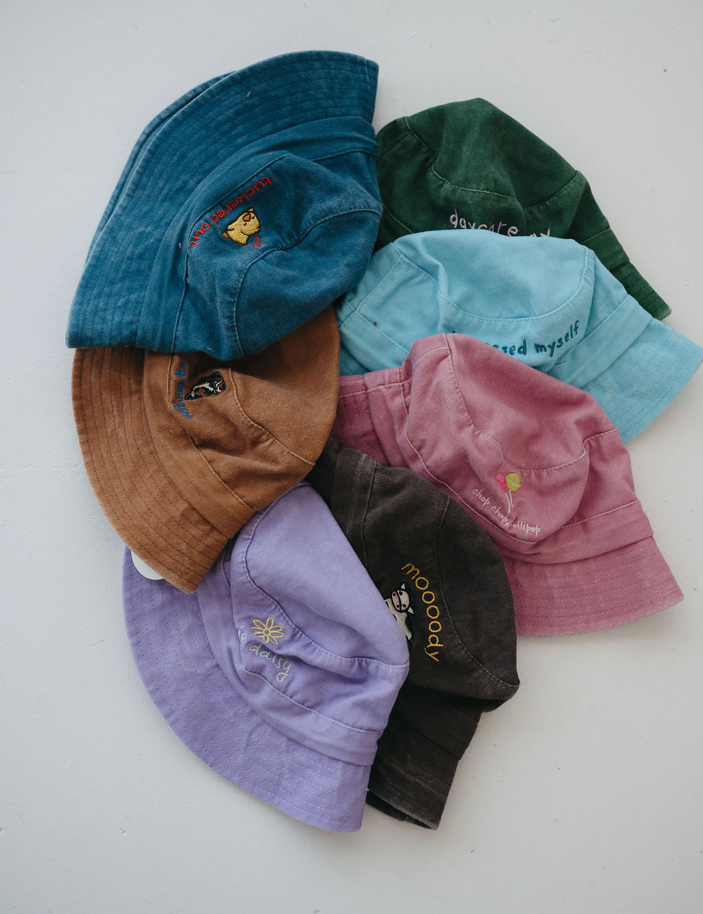 A group of seven toddler daycare washed cotton bucket hats in various colours (light blue, green, brown, dark blue, pink, purple, dark brown) arranged in a circle on a plain white background.