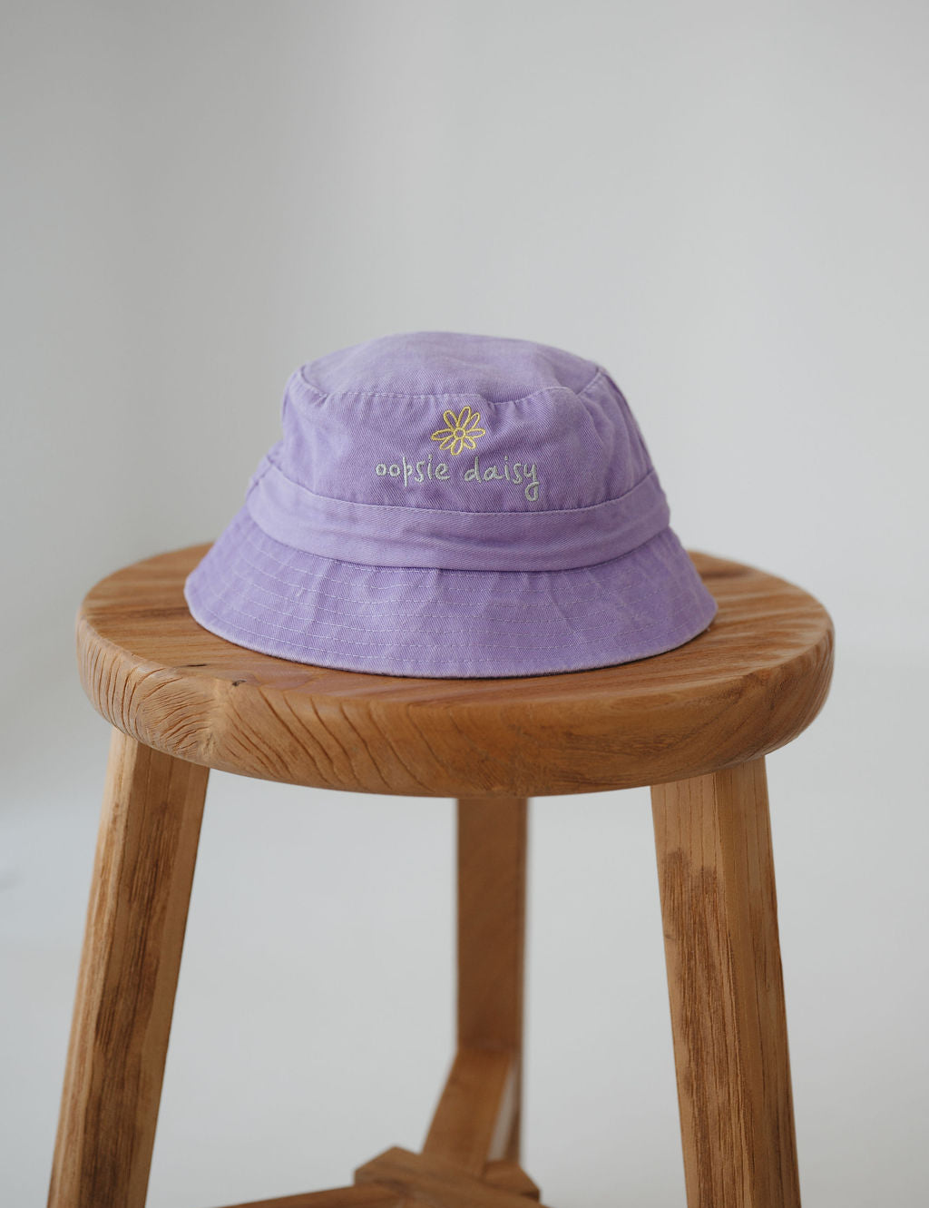 A light purple/ lavender washed cotton toddler daycare bucket hat with a small embroidered yellow daisy and white text reading 'oopsie daisy' on the front, displayed on a round wooden stool against a white background.