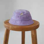 A light purple/ lavender washed cotton toddler daycare bucket hat with a small embroidered yellow daisy and white text reading 'oopsie daisy' on the front, displayed on a round wooden stool against a white background.