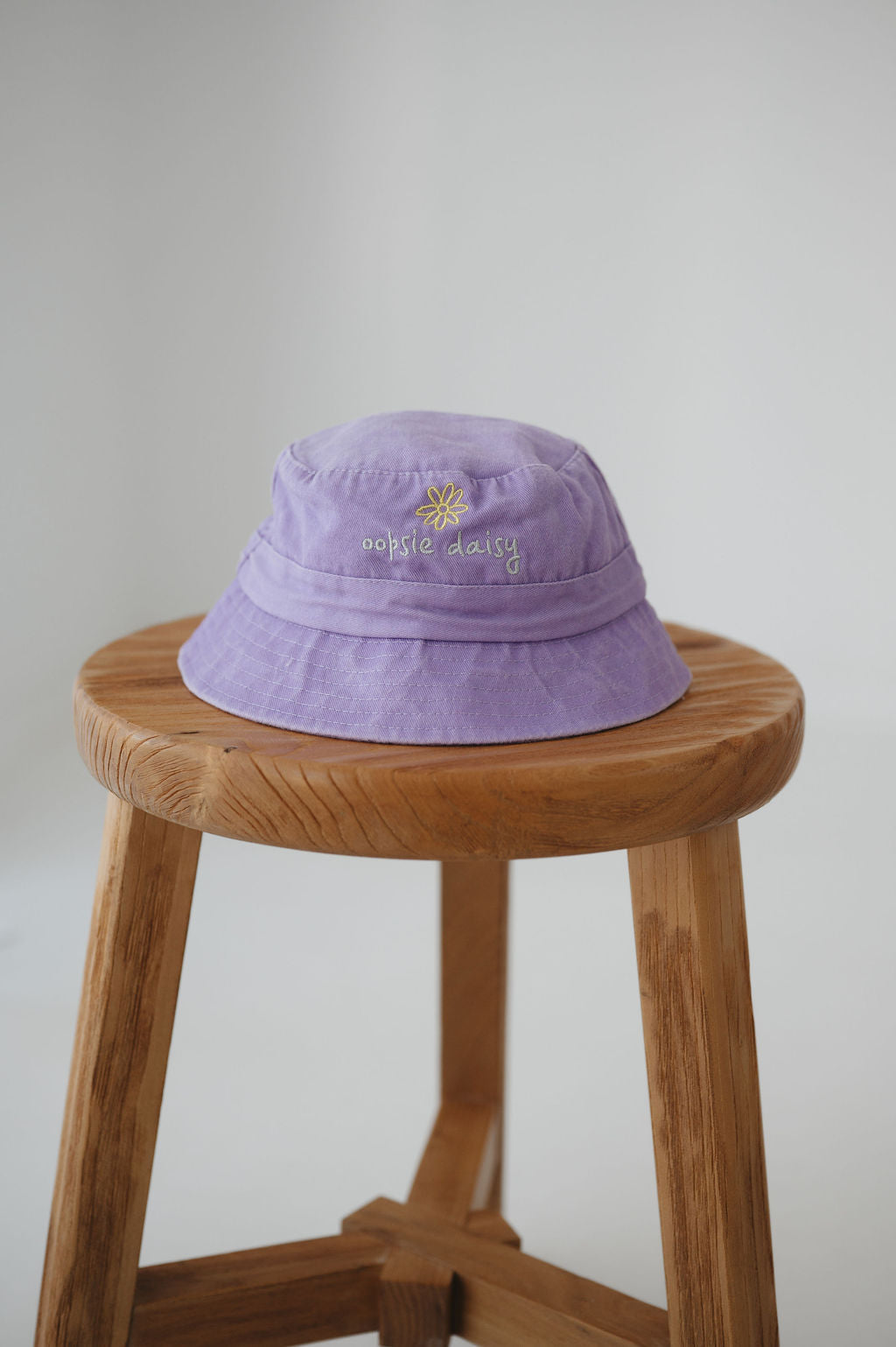 A light purple/ lavender washed cotton toddler daycare bucket hat with a small embroidered yellow daisy and white text reading 'oopsie daisy' on the front, displayed on a round wooden stool against a white background.