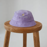 A light purple/ lavender washed cotton toddler daycare bucket hat with a small embroidered yellow daisy and white text reading 'oopsie daisy' on the front, displayed on a round wooden stool against a white background.