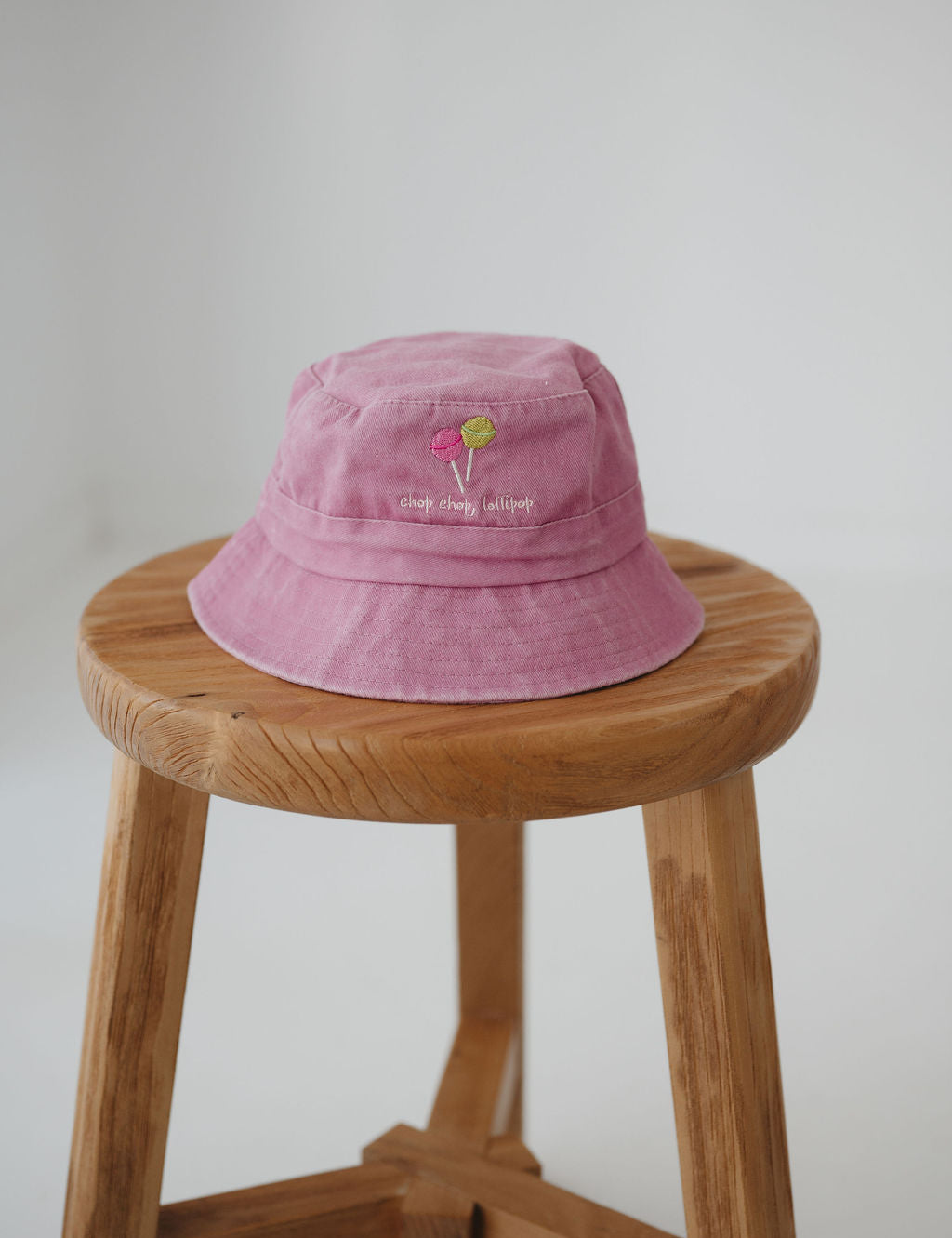 A pink bucket hat with two small embroidered lollipop graphic with the phrase "chop, chop lollipop." underneath. The toddler daycare bucket hat sits on a round wooden stool against a plain light white background.