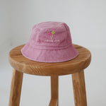 A pink bucket hat with two small embroidered lollipop graphic with the phrase "chop, chop lollipop." underneath. The toddler daycare bucket hat sits on a round wooden stool against a plain light white background.