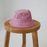 A pink bucket hat with two small embroidered lollipop graphic with the phrase "chop, chop lollipop." underneath. The toddler daycare bucket hat sits on a round wooden stool against a plain light white background.