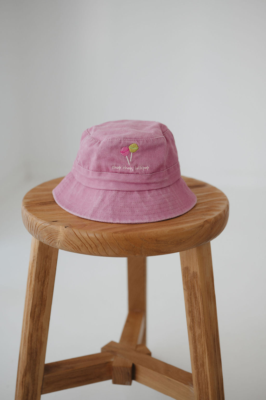 A pink bucket hat with two small embroidered lollipop graphic with the phrase "chop, chop lollipop." underneath. The toddler daycare bucket hat sits on a round wooden stool against a plain light white background.