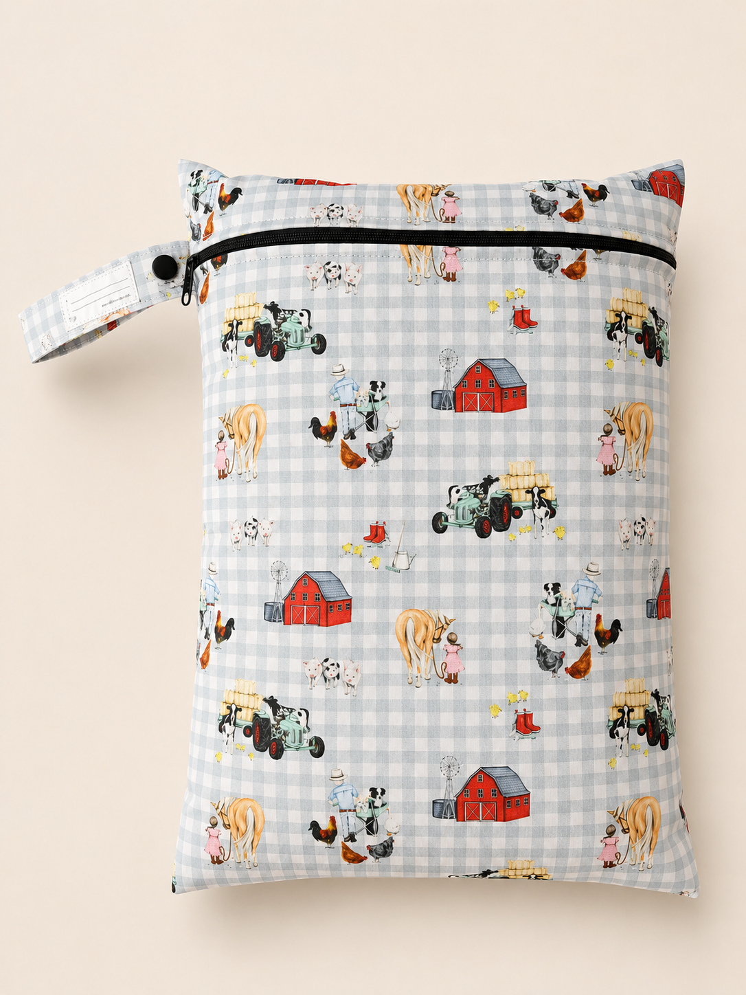 A rectangle PUL fabric daycare wet bag  with a zipper closure and a button loop strap, printed with a hand sketched farm-themed pattern on a light blue and white gingham background. The print includes red barns, tractors, cows, chickens, and other farm animals with a girl and a horse and a boy with his dog. Shown against a solid light beige background.