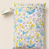 A PUL material zippered wet bag with a colourful hand sketched floral print on a cream background, featuring a matching fabric wrist strap with button closure, shown against a solid light beige background. Floral colours a soft purple, soft pink and soft yellow with soft green stems.