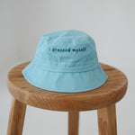 A blue washed cotton toddler daycare bucket hat with "I dressed myself" embroidered in a dark blue text on the front, displayed on a round wooden stool against a white background.