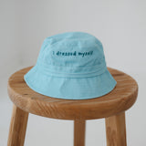 A blue washed cotton toddler daycare bucket hat with "I dressed myself" embroidered in a dark blue text on the front, displayed on a round wooden stool against a white background.