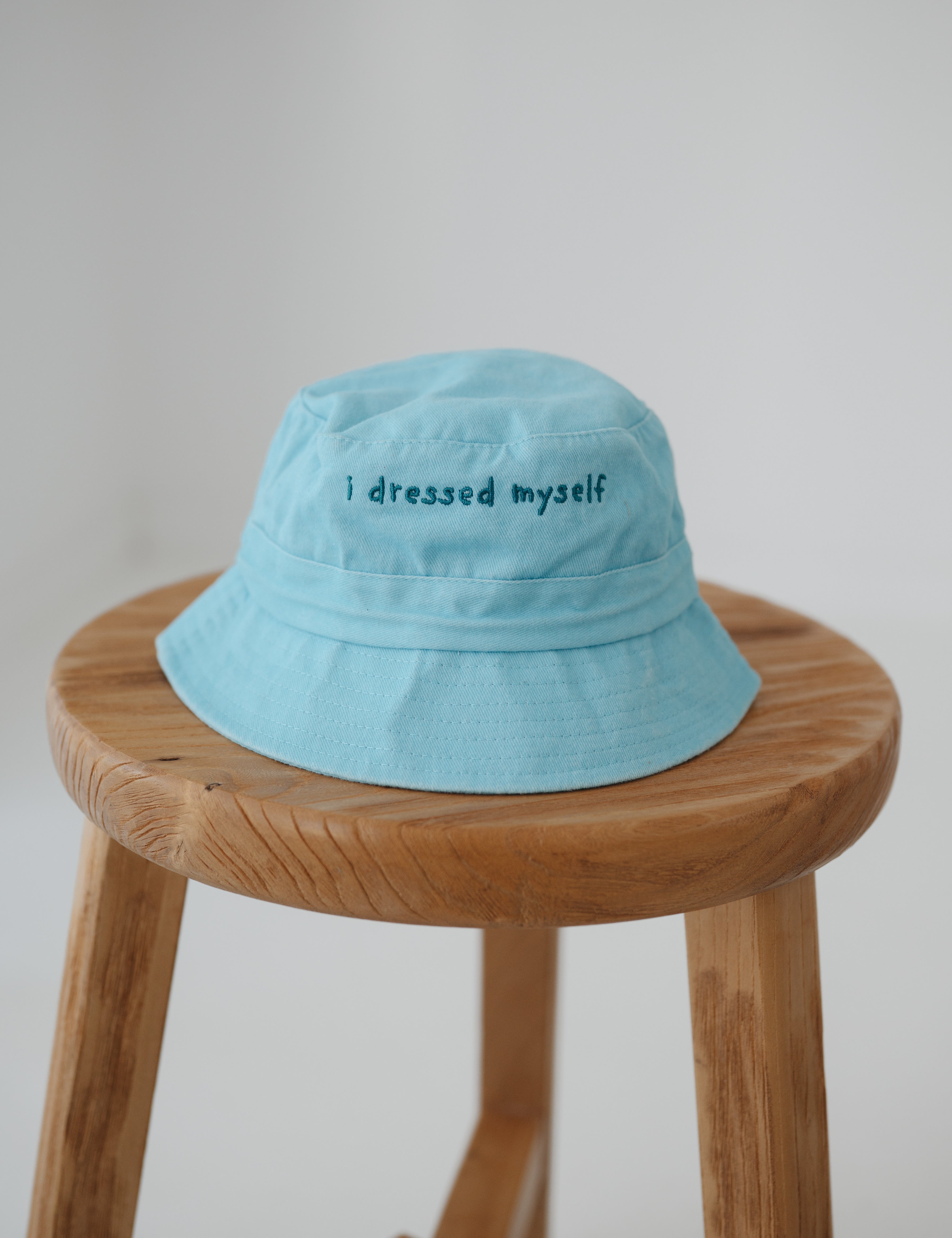 A blue washed cotton toddler daycare bucket hat with "I dressed myself" embroidered in a dark blue text on the front, displayed on a round wooden stool against a white background.