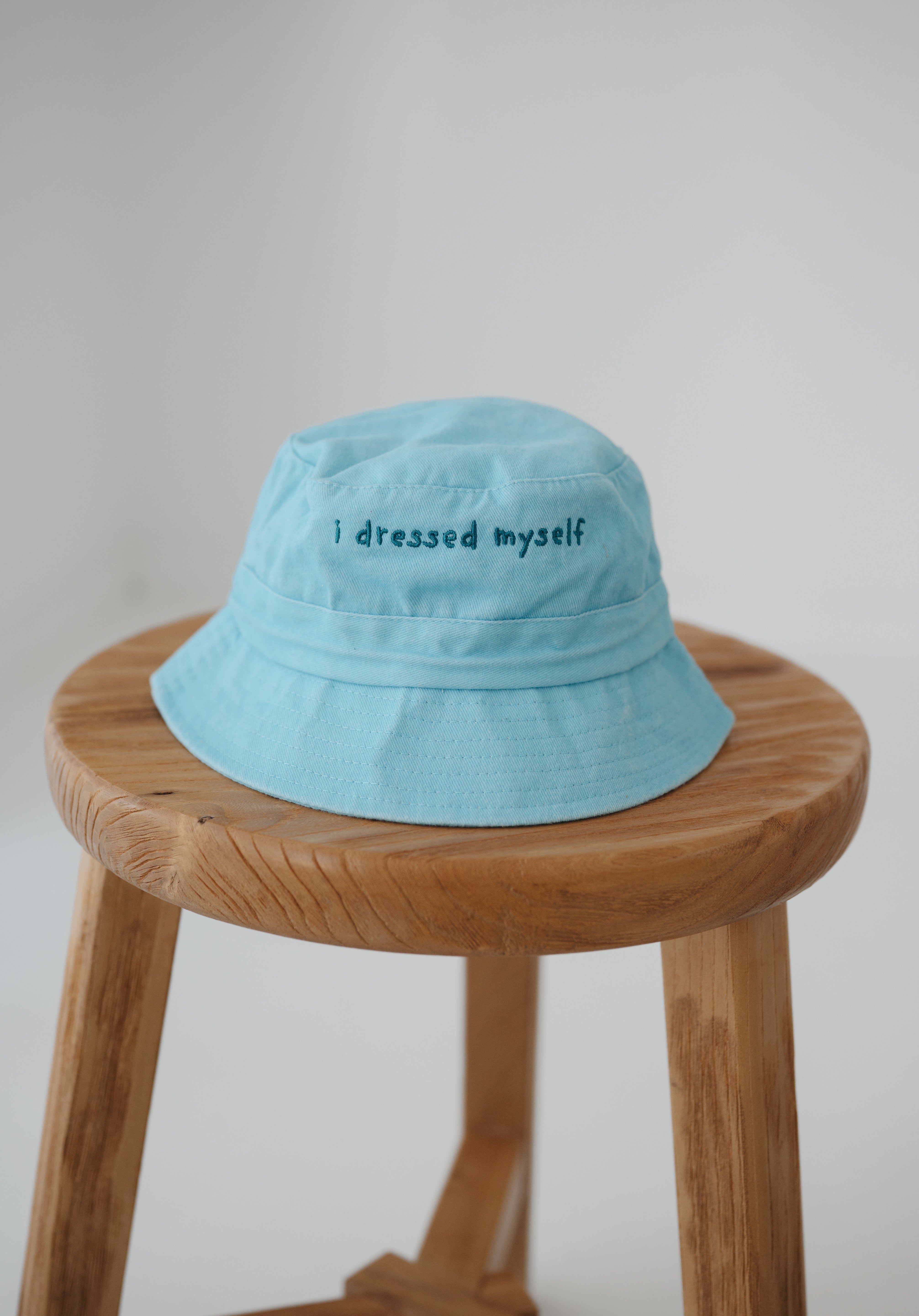 A blue washed cotton toddler daycare bucket hat with "I dressed myself" embroidered in a dark blue text on the front, displayed on a round wooden stool against a white background.